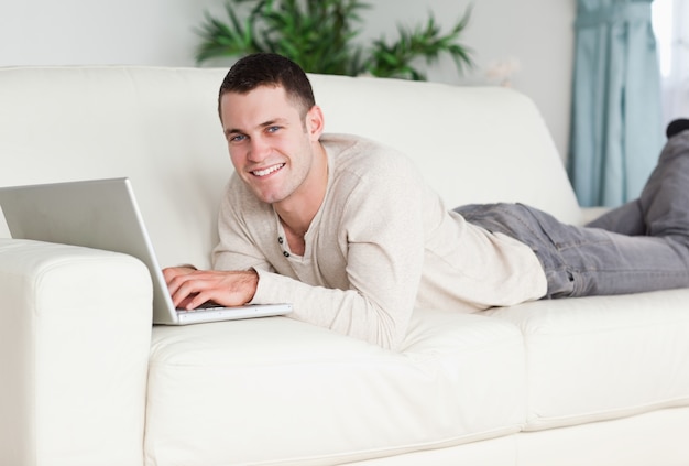 Sonriente hombre acostado en un sofá con una computadora portátil