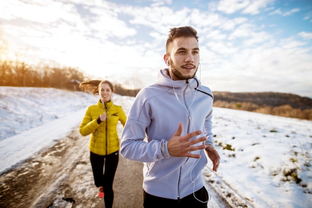 Sonriente hombre activo en ropa deportiva de invierno con auriculares con una niña sonriente con una cola de caballo afuera en la nieve