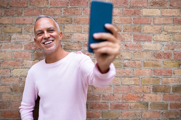 Sonriente hombre adulto caucásico tomando selfie. Fondo de pared de ladrillo naranja. Copie el espacio. Concepto de tecnología y estilo de vida.