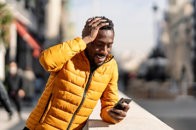 Sonriente hombre africano confundido con teléfono móvil quejándose de error sentado en una ciudad