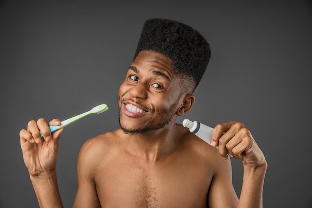 Sonriente hombre africano medio desnudo sosteniendo cepillo de dientes y pasta de dientes aislado sobre fondo gris