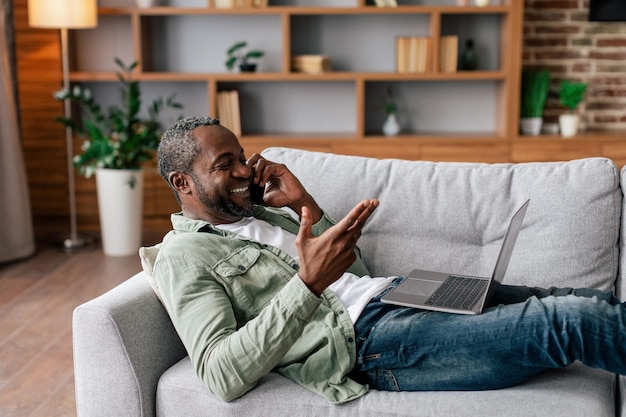 Sonriente hombre afroamericano de mediana edad en llamadas casuales por trabajo telefónico con computadora portátil sentado en el sofá