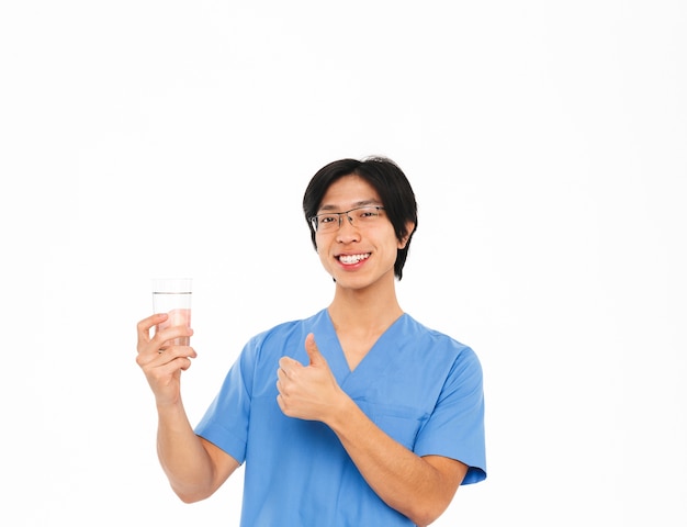 Sonriente hombre asiático médico con uniforme que se encuentran aisladas sobre una pared blanca, mostrando el vaso con agua, pulgares arriba