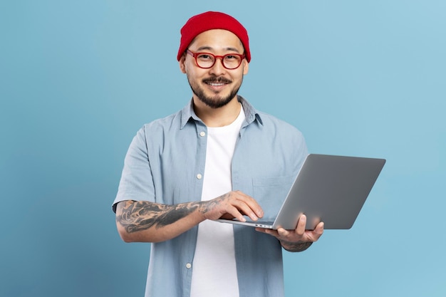 Sonriente hombre asiático programador independiente sosteniendo un ordenador portátil aislado sobre fondo azul.