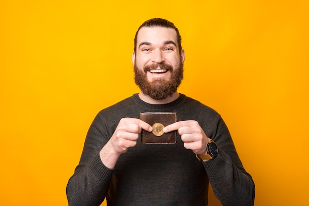 Sonriente hombre barbudo de pie sobre una pared amarilla y mostrando bitcoin cerca de la billetera