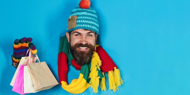 Foto sonriente hombre barbudo en ropa de invierno con bolsas de compras mirando a través de un agujero de papel hombre guapo en