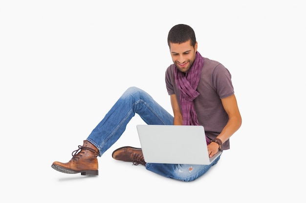 Sonriente hombre con bufanda sentada en el piso usando laptop