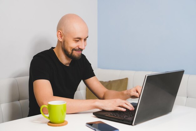 Sonriente hombre Calvo con barba en camiseta negra trabajando en su portátil en casa vista lateral