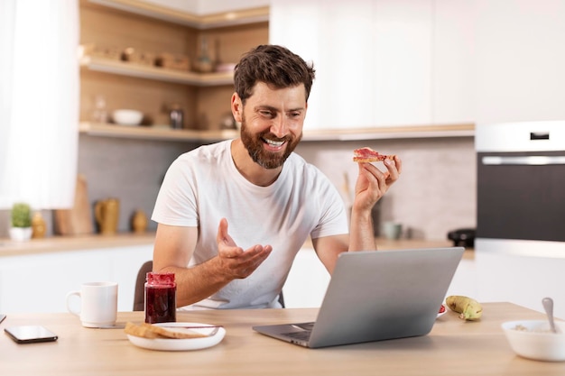 Sonriente hombre caucásico de mediana edad con barba en camiseta blanca come sándwich tiene videollamada gesticula