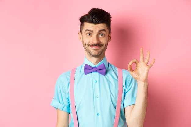 Sonriente hombre caucásico con pajarita y tirantes mostrando todos los gestos bien, recomendando algo genial, alabando la promoción sobre fondo rosa.