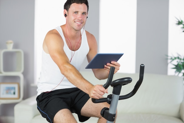 Sonriente hombre deportivo haciendo ejercicio en bicicleta y sosteniendo la tableta