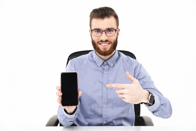 Sonriente hombre elegante barbudo en anteojos que muestra la pantalla del teléfono inteligente en blanco y apuntando mientras está sentado junto a la mesa