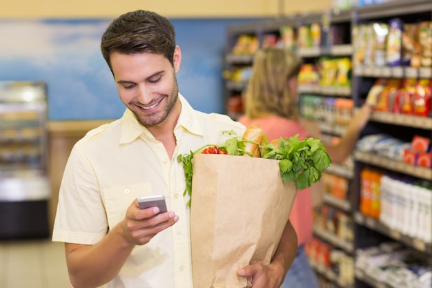 Sonriente hombre guapo comprar productos alimenticios y usar su teléfono inteligente