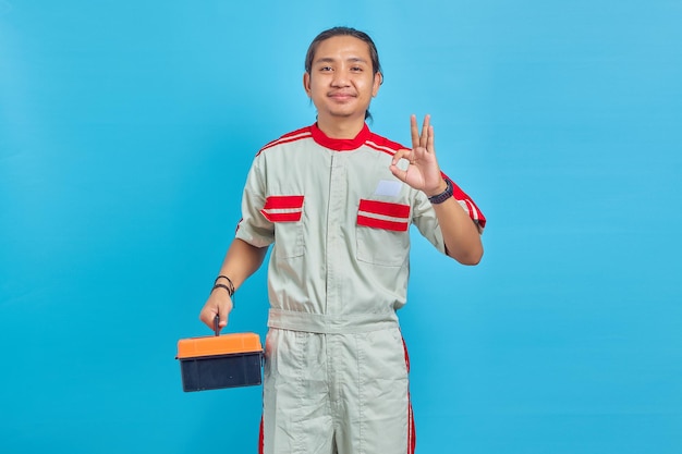 Sonriente hombre guapo vestido con uniforme de mecánico sosteniendo la caja de herramientas y mostrando gesticular signo bien