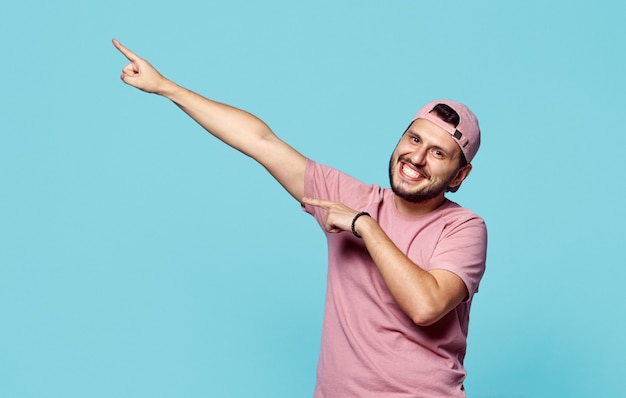 Sonriente hombre hipster barbudo apuntando a un lado en la pared azul colorida