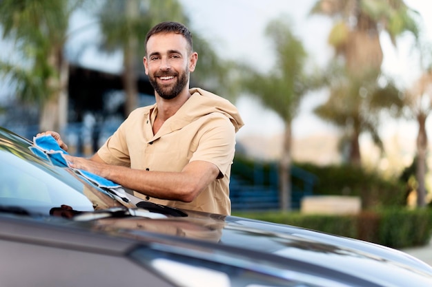 Sonriente hombre hispano confiado limpiando auto usando tela de microfibra. Concepto de servicio de lavado de autos