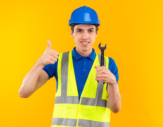 Sonriente a hombre joven constructor en uniforme sosteniendo una llave de boca que muestra el pulgar hacia arriba aislado en la pared amarilla