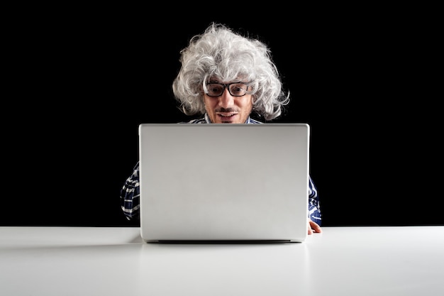 Sonriente hombre mayor con cabello gris vistiendo una camisa a cuadros usando laptop sentarse en el escritorio. Fondo negro con espacio de copia