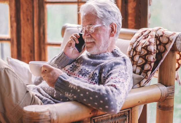 Sonriente hombre mayor de pelo blanco en suéter de invierno sentado en la sala de estar sosteniendo una taza de té mediante teléfono móvil Abuelo anciano despreocupado disfrutando de tecnología y redes sociales. Chalet rústico en el bosque