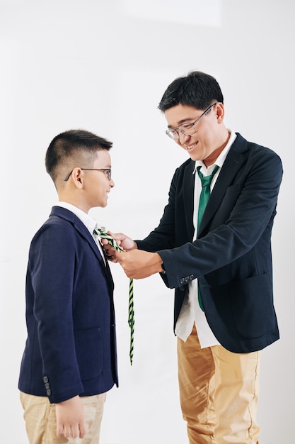 Sonriente hombre de mediana edad asiático ayudando a su hijo a ajustar la corbata cuando se prepara para la celebración
