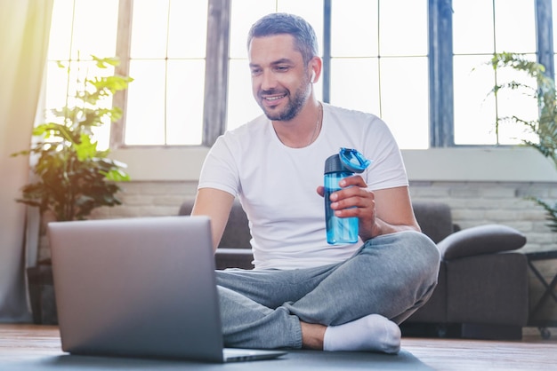 Sonriente hombre de mediana edad usando laptop y bebiendo agua mientras toma un descanso durante el entrenamiento en casa