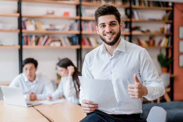 Sonriente hombre del Medio Oriente trabajando en la oficina diversa y mostrando los pulgares para arriba