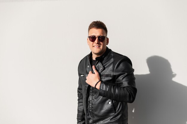 Sonriente hombre de moda brutal con elegantes gafas de sol negras y una moderna chaqueta de cuero negro afuera cerca de una pared gris en un día soleado