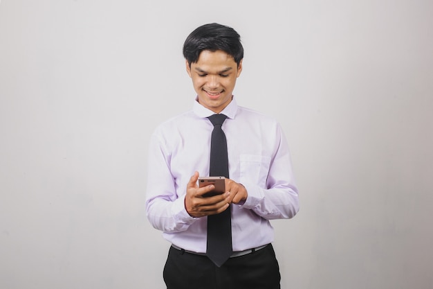 Sonriente hombre de negocios asiático en camisa blanca y corbata negra de pie y usando teléfonos inteligentes.