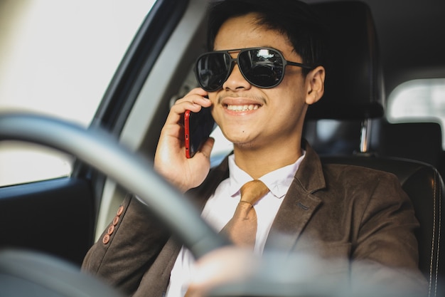 Sonriente hombre de negocios asiático con gafas de sol está conduciendo un coche mientras conversa en un teléfono inteligente.