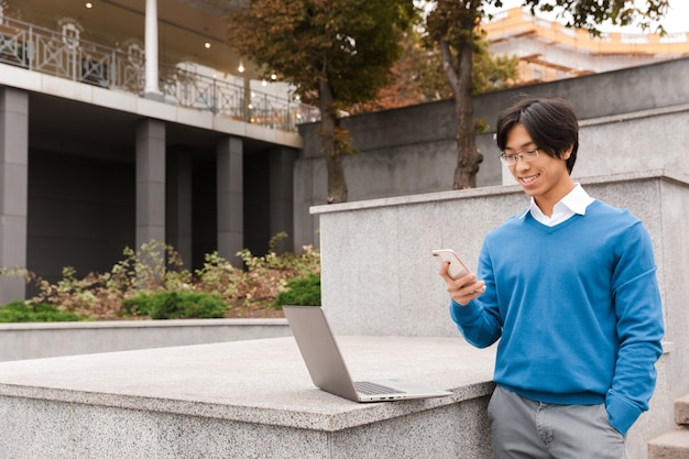Sonriente hombre de negocios asiático de pie al aire libre con ordenador portátil y teléfono móvil