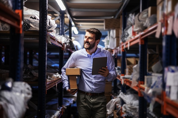 Sonriente hombre de negocios barbudo caminando a través del almacenamiento de la empresa de transporte, sosteniendo cajas debajo de la axila y usando la tableta para verificar las mercancías.