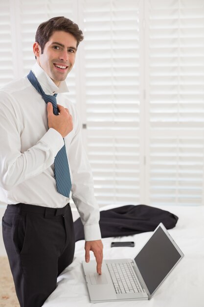 Sonriente hombre de negocios con corbata mientras usa la computadora portátil en la habitación del hotel