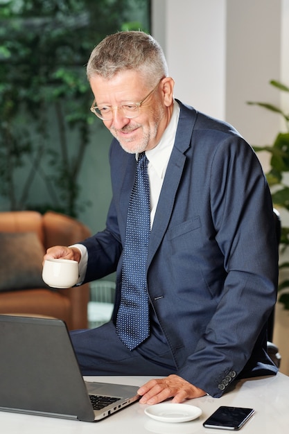 Sonriente hombre de negocios de edad tomando café por la mañana y leyendo correos electrónicos en la pantalla del portátil