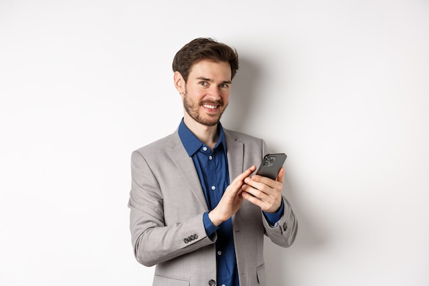 Sonriente hombre de negocios exitoso con smartphone sobre fondo blanco, de pie en traje.