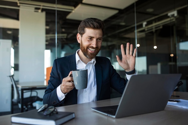 Sonriente hombre de negocios de mediana edad agitando la mano en la cámara web de la computadora portátil que se reúne en el espacio libre interior de la oficina