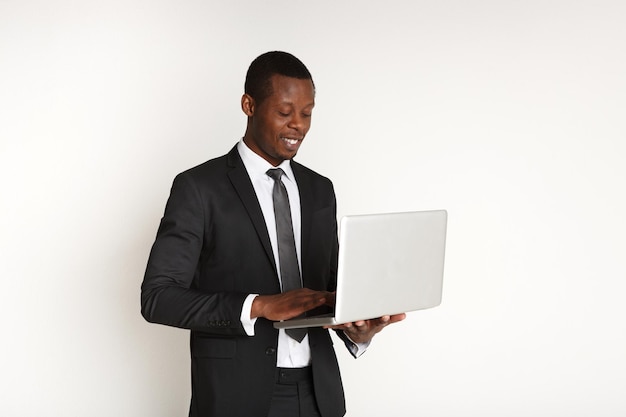 Sonriente hombre de negocios negro con portátil aislado. Hombre joven en traje trabajando en equipo mientras está de pie, fondo blanco. Dispositivo portátil. comercio electrónico, concepto de negocio