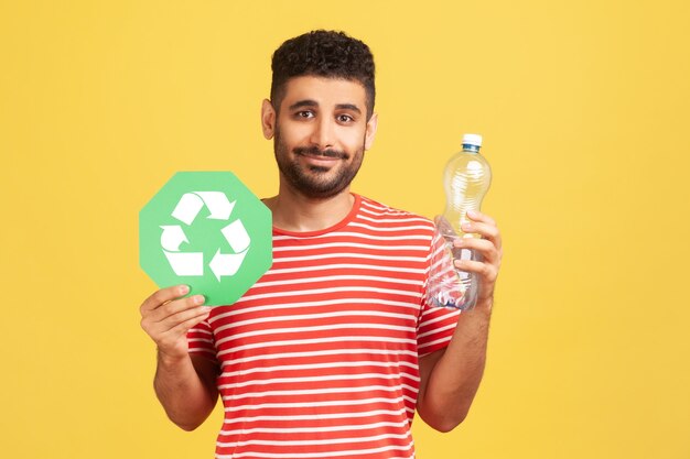 Sonriente hombre positivo con barba en camiseta a rayas sosteniendo una botella de plástico y reciclando el símbolo verde, clasificando su basura, salvando la ecología. Tiro de estudio interior aislado sobre fondo amarillo