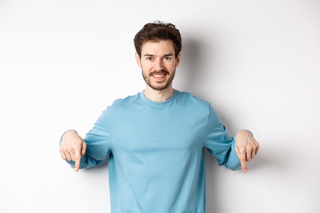 Sonriente hombre seguro apuntando con el dedo hacia abajo, mostrando un banner o logotipo promocional sobre fondo blanco, de pie en una sudadera azul casual