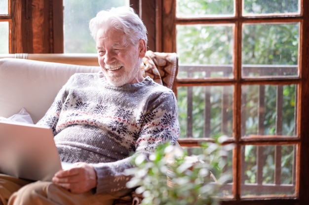 Sonriente hombre senior de pelo blanco en suéter de invierno sentado en la sala de estar usando la computadora portátil. Abuelo mayor despreocupado disfrutando de la tecnología y las redes sociales. Chalet rústico en el bosque