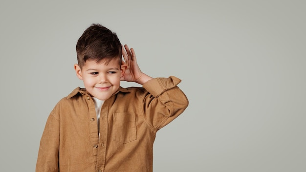 Sonriente interesado inquisitivo pequeño niño europeo presiona la mano al oído escucha buenas noticias