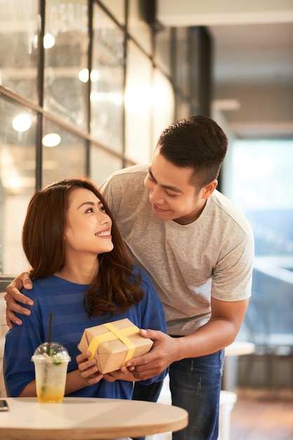 Foto sonriente joven abrazando a la novia asiática desde atrás en el café y dándole un regalo