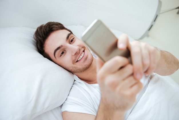 Sonriente joven acostado y usando un teléfono móvil en la cama
