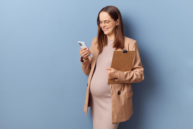 Sonriente joven adulta embarazada mujer de negocios con vestido y chaqueta posando aislada sobre un fondo azul sosteniendo el portapapeles y usando el teléfono móvil para charlar profesionalmente