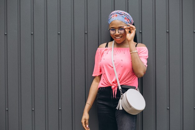 sonriente joven africana en unas gafas con bolsa posando contra la pared gris
