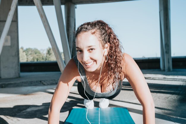 Sonriente joven africana practicando yoga, estirando las piernas sobre la colchoneta, instructor de salud. Hacer ejercicio, vistiendo ropa deportiva, pantalón negro y top, ciudad urbana al aire libre en toda su longitud, mirando a la cámara