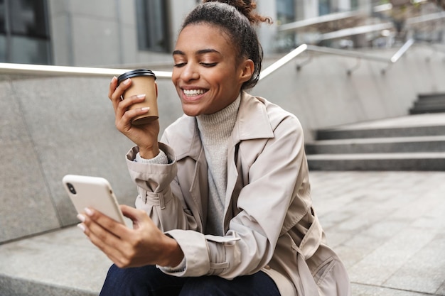 Sonriente joven africana vistiendo abrigo de otoño sentado en una escalera en la calle, mediante teléfono móvil mientras bebe café para llevar