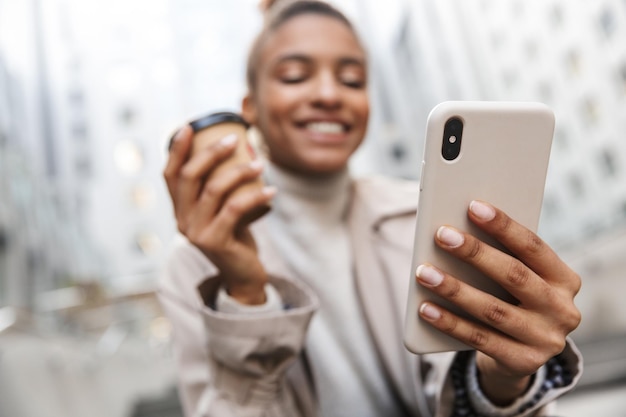 Sonriente joven africana vistiendo abrigo de otoño sentado en una escalera en la calle, mediante teléfono móvil mientras bebe café para llevar