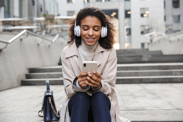 Sonriente joven africana vistiendo abrigo de otoño sentado en una escalera en la calle, usando el teléfono móvil mientras escucha música con auriculares inalámbricos