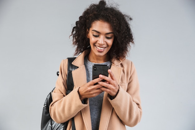 Sonriente joven africana vistiendo abrigo mediante teléfono móvil mientras se encuentra aislado