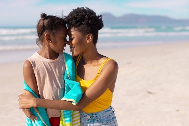 Sonriente joven afroamericana madre y niña con cara a cara en la playa contra el mar en un día soleado